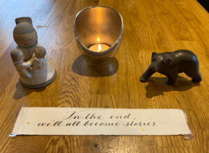 Image shows a pottery figurine of an Isleta storyteller, a candle, and a bear figurine. Below is a quote from Margaret Atwood, 'In the end, we'll all become stories.'